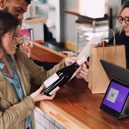 A cheerful woman at a retail counter holds a wine bottle while interacting with a cashier. A point-of-sale tablet with a QR code is visible on the counter, and another person stands beside her holding a shopping bag.