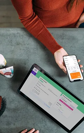 A person in a rust-colored sweater holds a smartphone displaying a payment screen, next to a tablet showing an online checkout summary on a gray countertop.