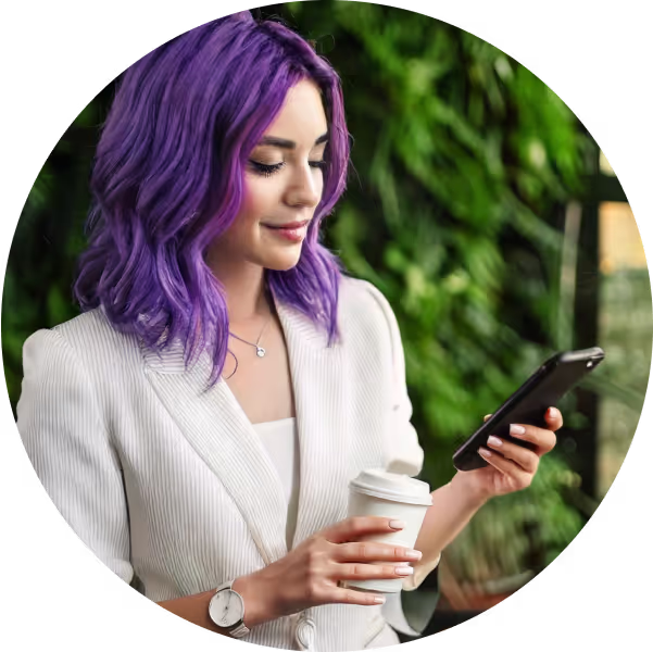 Smiling woman with vibrant purple hair, wearing a light pinstripe blazer, holding a coffee cup in one hand and a smartphone in the other. She is standing in front of a lush green vertical garden, creating a fresh and modern atmosphere.